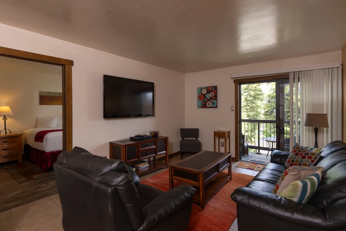 The image shows a cozy living room with a black leather sofa set adorned with colorful pillows, a wooden coffee table, and a flat screen TV mounted on the wall. Sunlight pours in through a sliding glass door that opens onto a balcony, offering a glimpse of mountain views and inviting guests to relax both indoors and out.