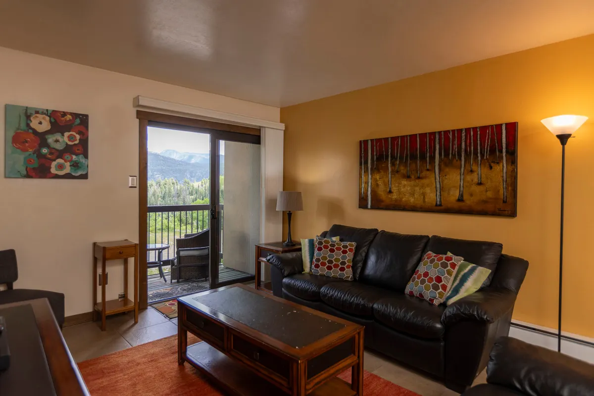A welcoming living room featuring a black leather sofa adorned with colorful pillows, a nature-themed wall painting, a wooden coffee table, and a sliding glass door that opens to a balcony with expansive mountain views. The room is filled with natural light and exudes a cozy, communal ambiance.