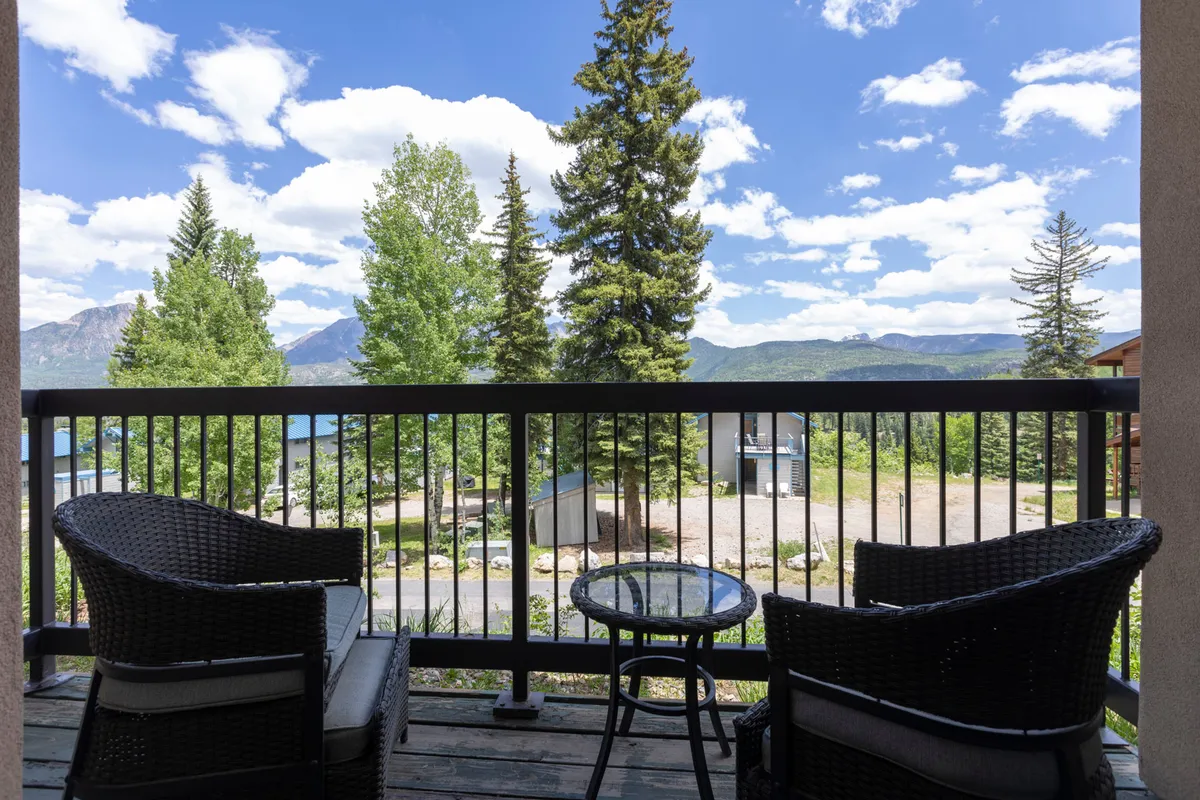 A cozy outdoor balcony furnished with wicker chairs and a glass-top table, framed by a black railing. The balcony overlooks lush green trees and a stunning mountain range, offering a serene space for guests to relax and enjoy the view after a day on the slopes.