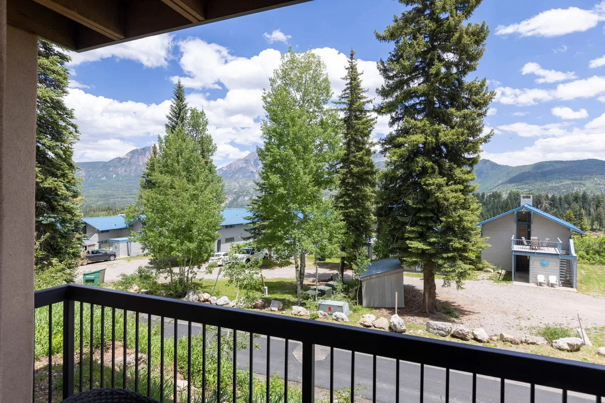 A balcony view framed by a black railing, overlooking tall conifer and deciduous trees, distant mountain peaks, and a blue-roofed lodge. The scene is tranquil, showcasing the direct connection between the accommodation and the surrounding natural beauty.