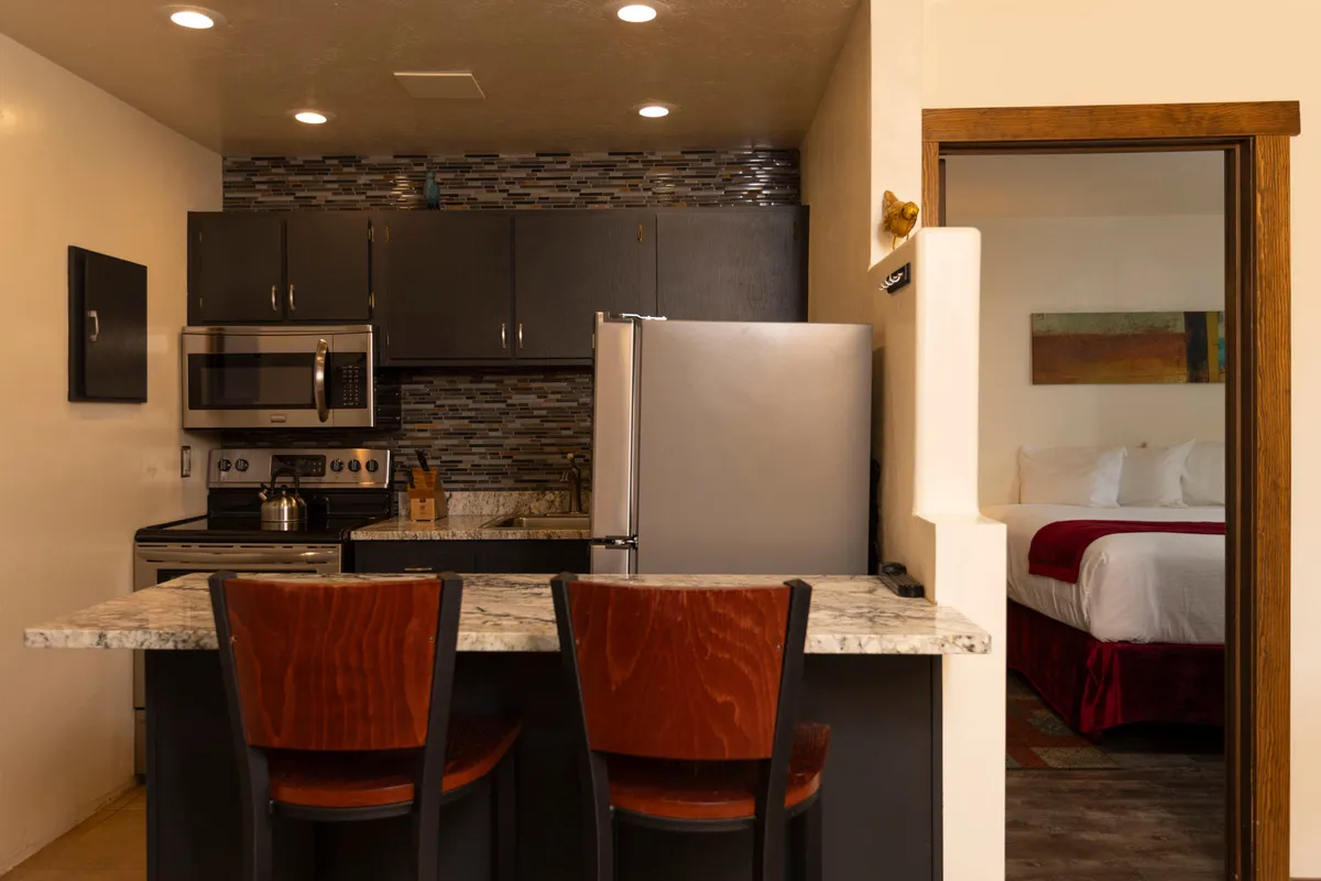 A warm, homey kitchen scene with a countertop for casual dining, two wooden chairs, a visible stainless steel refrigerator, stove, and microwave. The open view into an adjacent bedroom adds a sense of connectivity and family comfort.