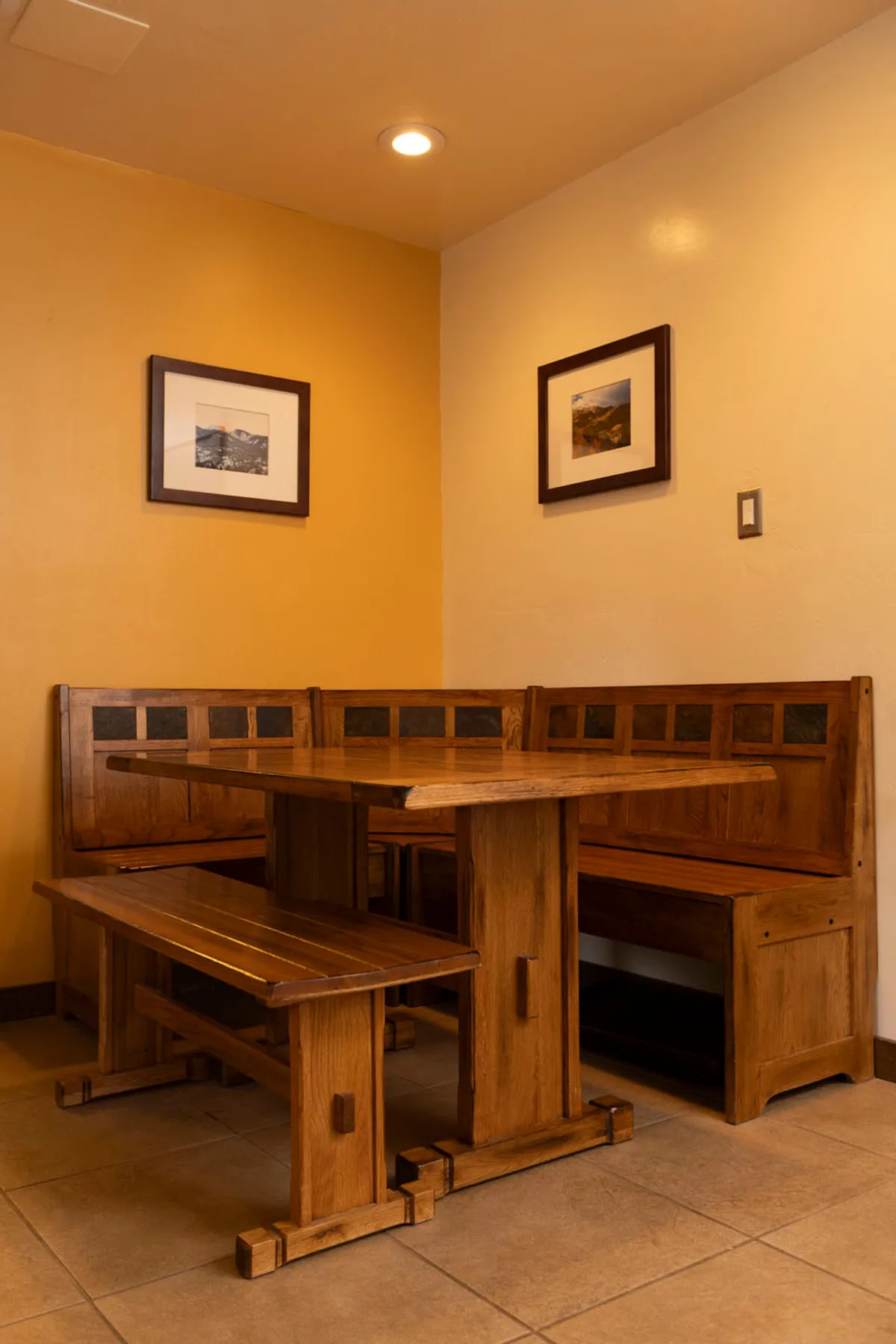 A cozy dining nook with solid wood corner bench seating, a sturdy wooden table, and simple framed landscape photographs on the walls. The setting is intimate, designed for group meals, games, and conversation.