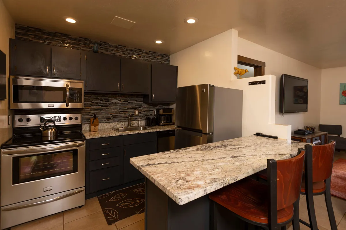 A modern kitchen with a granite countertop breakfast bar and wooden stools, stainless steel appliances, dark cabinetry with a mosaic tile backsplash, and an open view into the living area with a mounted TV. The setup encourages socializing while preparing food.