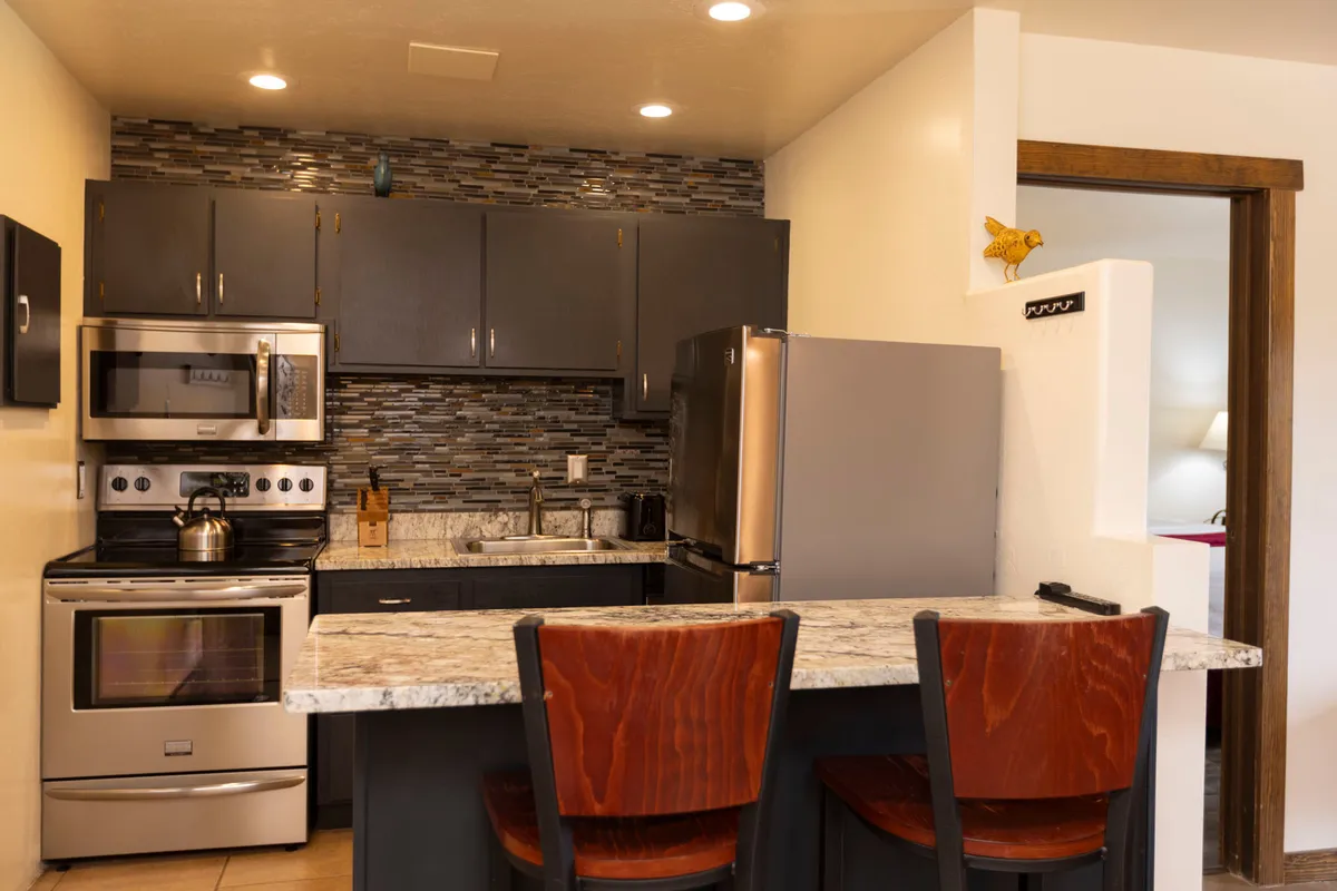 A modern and inviting kitchen featuring a marble countertop island with wooden chairs, stainless steel appliances including an oven and refrigerator, dark cabinetry with a mosaic tile backsplash, and a well-equipped sink area with utensils. The setup clearly showcases ample counter space and full-sized appliances.