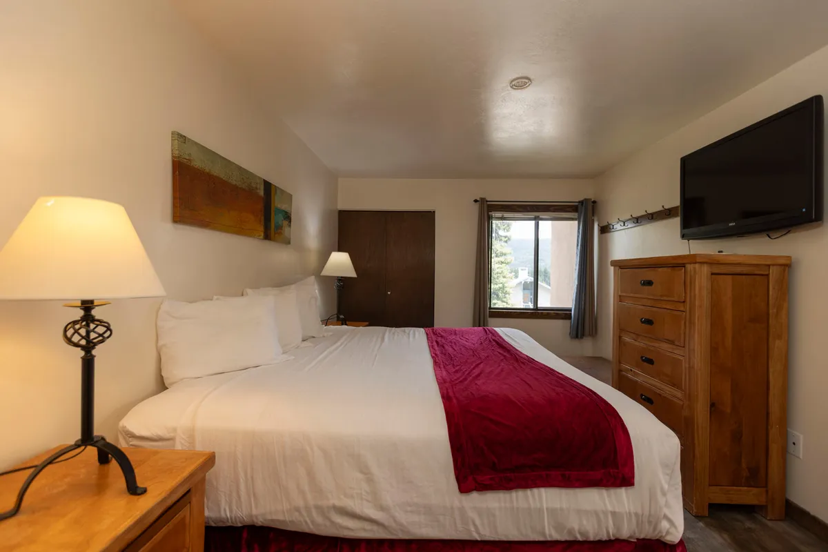 The image displays the master bedroom, featuring a plush king bed adorned with crisp white linens and a red throw. A wooden dresser with a mounted TV sits opposite the bed, while soft bedside lamps create a warm, inviting glow. The window provides a glimpse of tranquil alpine scenery outside, reinforcing a restful atmosphere.
