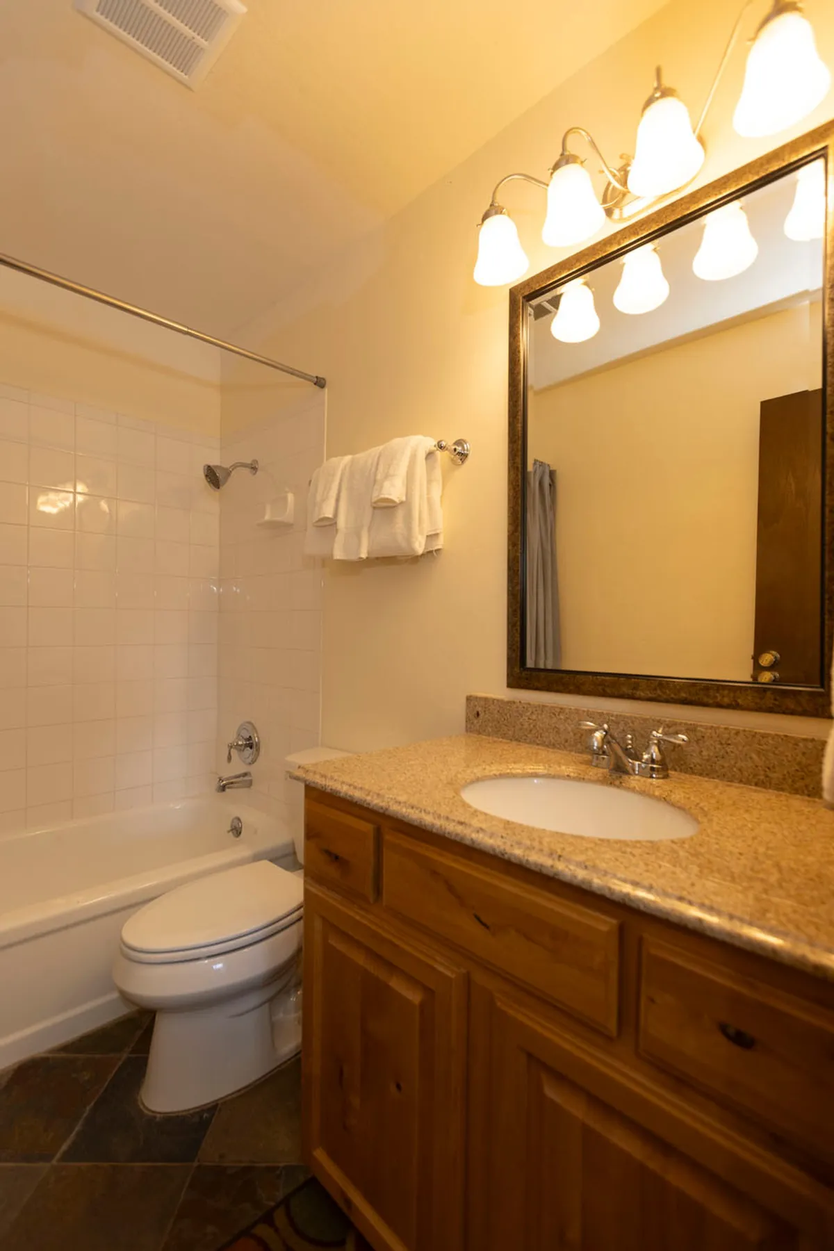 The photo shows a well-appointed bathroom with a bathtub and shower, wooden vanity with granite countertop and sink, large mirror with overhead lights, and toilet. The space is clean, practical, and inviting—ideal for families winding down after mountain adventures.