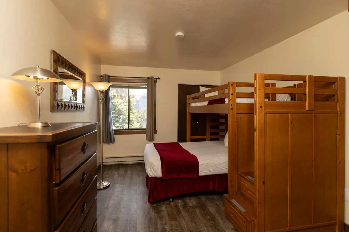 This image features the kid-friendly second bedroom with a sturdy wooden bunk bed—double bed below, single above—alongside a window offering mountain views. A dresser and floor lamp contribute to a cozy, family-friendly feel, ideal for children or extra guests.