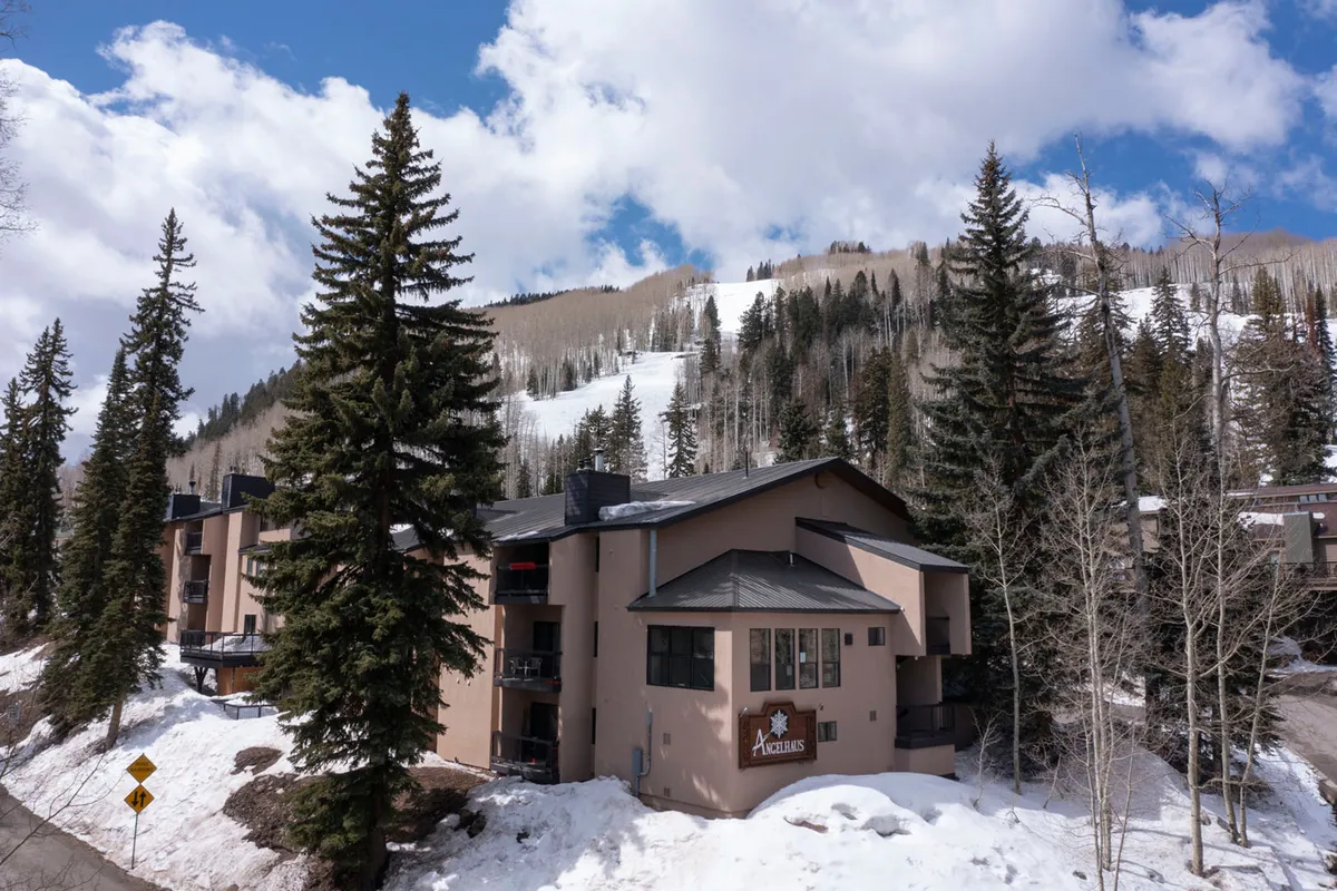This image shows the Angel Haus building with clear signage, set against a backdrop of snow-covered slopes and tall pine trees. The blue sky with scattered clouds enhances the lively alpine atmosphere, and the proximity of the property to the ski area is visually evident.