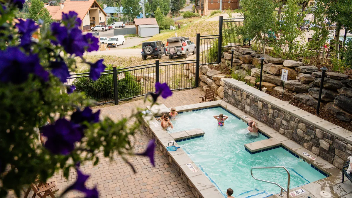 A smaller, heated community pool set amid natural stone landscaping and colorful purple flowers. People are seen enjoying the pool, creating a joyful and leisurely scene. The pool area is surrounded by natural beauty, blending relaxation with the mountain setting.