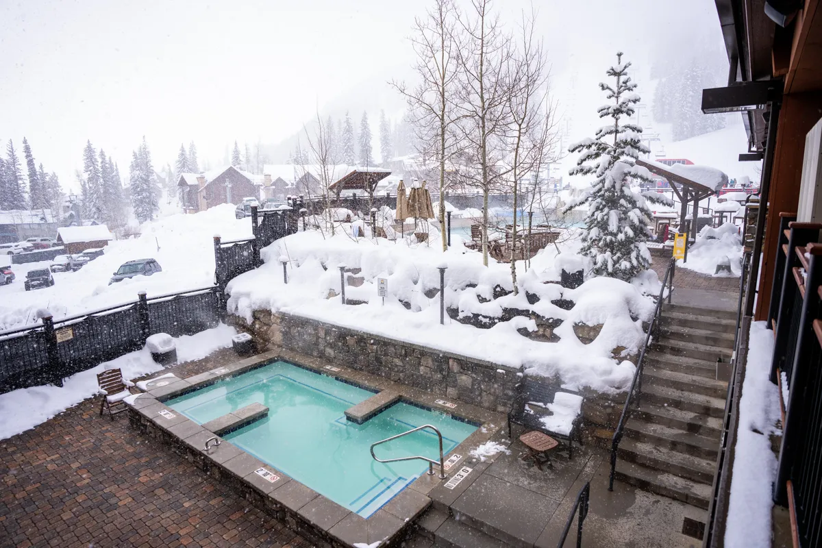 An inviting heated outdoor pool surrounded by a snowy landscape, with a stone retaining wall and stairs leading up to the pool area. The nearby resort buildings and evergreen trees frame the scene, while steam rises from the water, emphasizing the contrast between the warm pool and the chilly mountain air. The atmosphere is peaceful and perfect for unwinding after outdoor adventure.