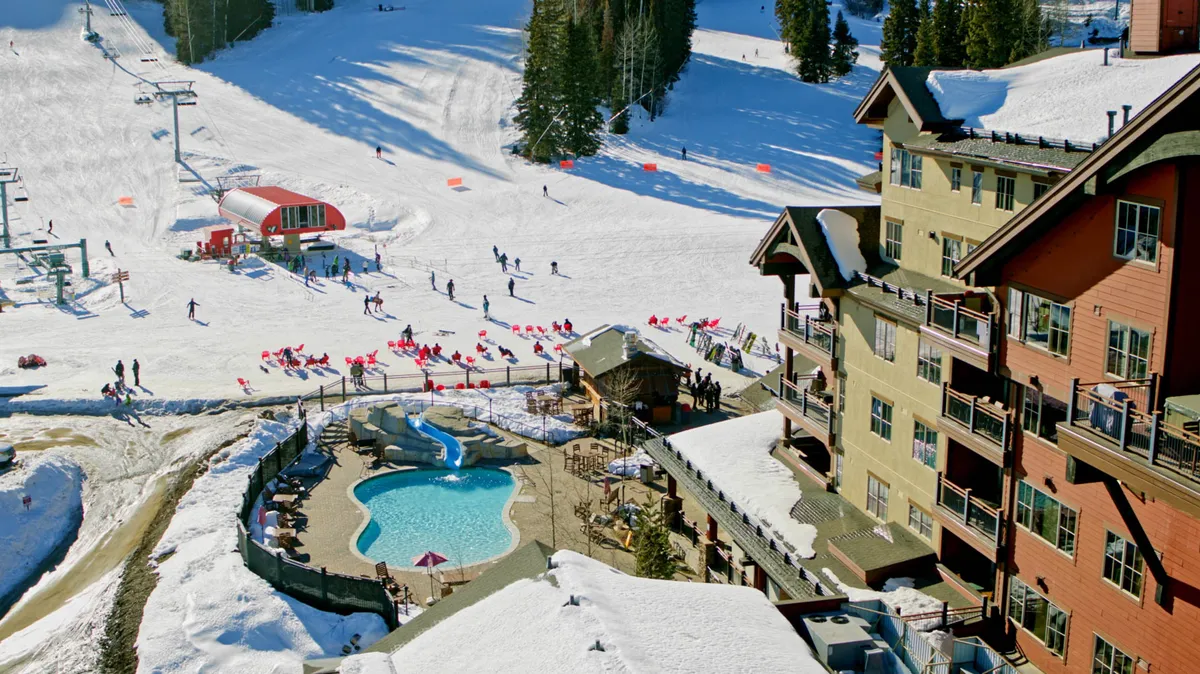 A vibrant exterior scene featuring a heated outdoor swimming pool with a water slide, surrounded by snow-covered ski slopes and a multi-story alpine lodge with balconies. Red lounge chairs are positioned near the pool area, offering a cozy retreat for sunbathing or relaxing after a day of skiing. The lively atmosphere is complemented by people enjoying both the pool and the wintry mountain surroundings.