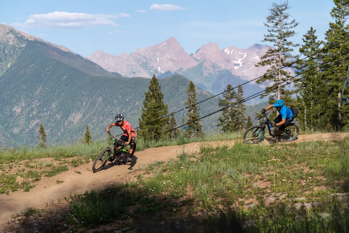 Two mountain bikers ride along a dirt trail that winds through lush forest and wildflowers, with a dramatic alpine mountain range towering in the background. The natural terrain and vibrant greenery speak to the area's outdoor recreational opportunities.