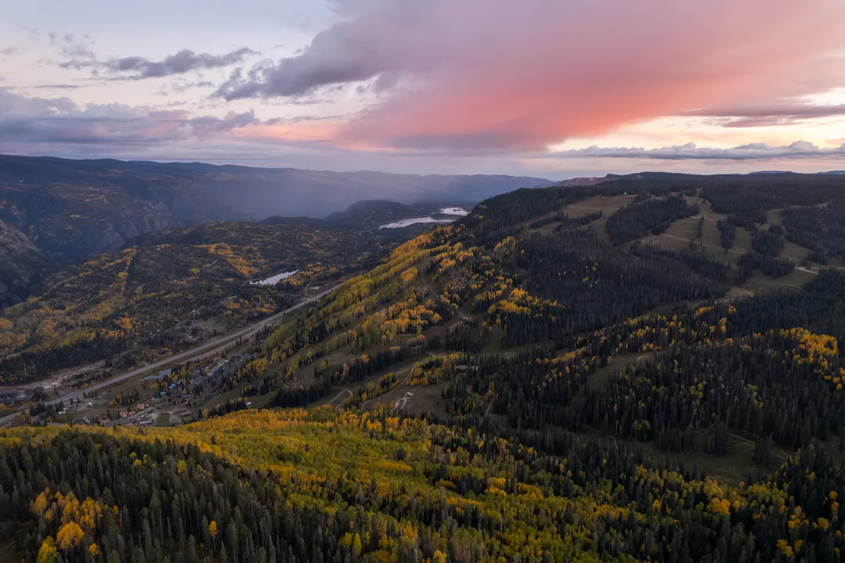 A breathtaking view of rolling hills and valleys blanketed in vibrant fall foliage, with mountain ridges fading into the distance beneath a soft sunset sky. A winding road traverses the landscape, highlighting the area's scenic beauty and outdoor appeal.