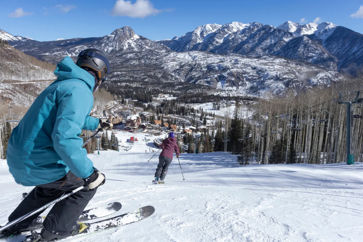 Two skiers descend a snowy slope with expansive alpine mountains in the background. The vibrant ski resort village and ski lift infrastructure are visible below, illustrating the dynamic winter sports environment and the lively community atmosphere.
