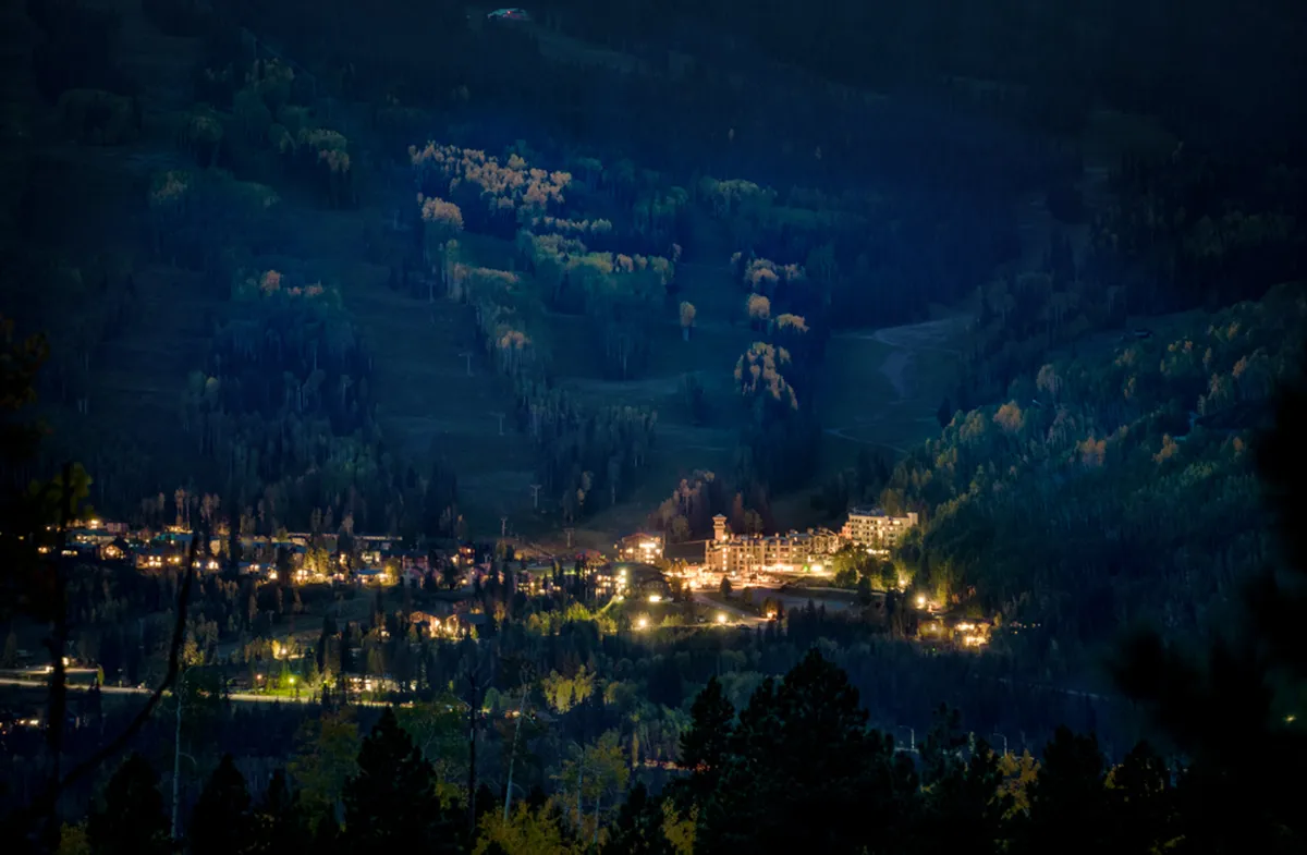 Resort buildings softly illuminated at night, nestled against a dark forested hillside, with scattered valley lights creating a cozy, serene atmosphere. The setting hints at peaceful evenings and the promise of returning to warm accommodations after a day outdoors.
